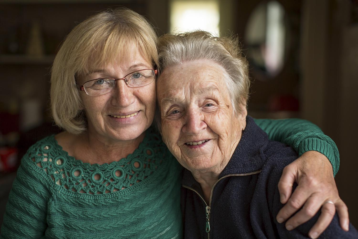 A mature woman has her arm around her aging mother as they smile at the camera.
