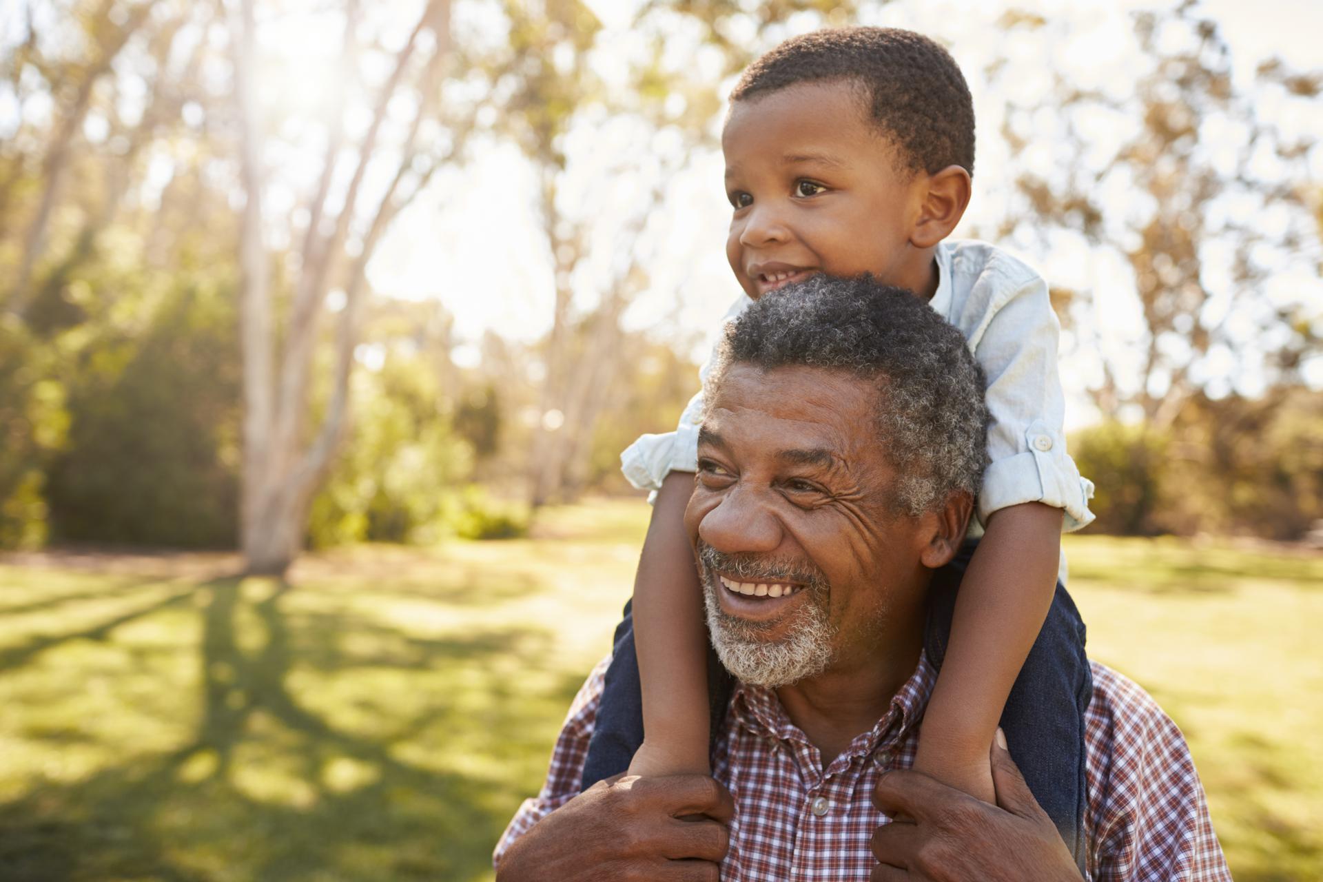 Black man with graying hair has a young child sitting on his shoulders outside in a park with a lot of trees and green grass.