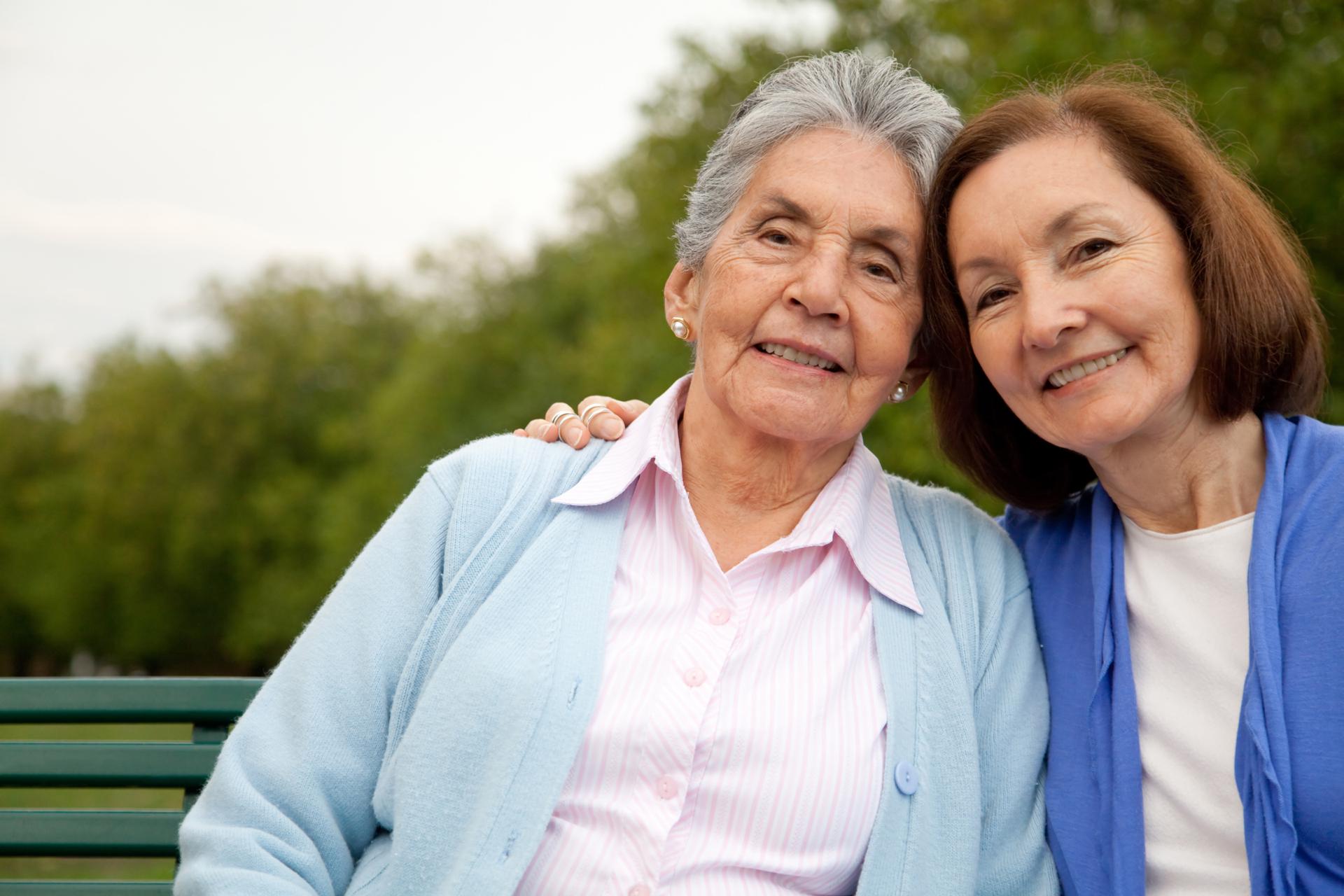 Older woman and mature daughter sit on a bench with their heads leaning in toward each other.